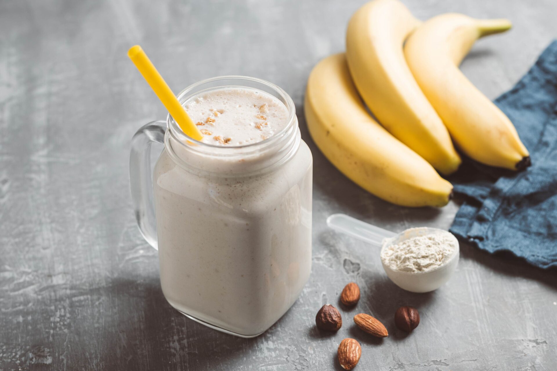 An Almond Protein Powder Smoothie next to a scoop of protein powder, bananas, and almonds - Protein Shakes and Smoothies